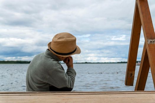 a boy in a hat sits on the shore of the lake and looks into the distance with his head propped on his hand, dreaming about something. cool weather, cloudy sky background. Summer holidays, vacations. rear view of a boy sitting on the shore of a lake and looking at the water