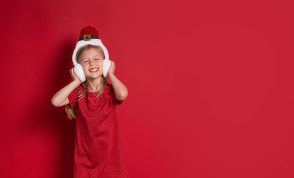 Long banner, red background with copy space. Merry Christmas and Happy New Year. Little blonde, caucasian woman in a santa claus hat, with headphones, laughs happily