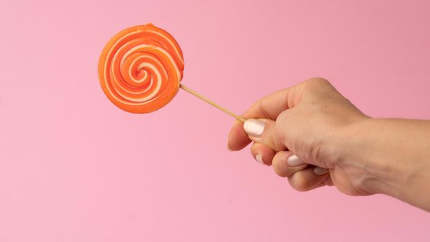 A woman's hand holds a multi-colored round lollipop on a pink background. High quality photo