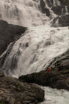 A woman wearing a red dress dancing next to Kjosfossen waterfall in a rocky landscape in Norway