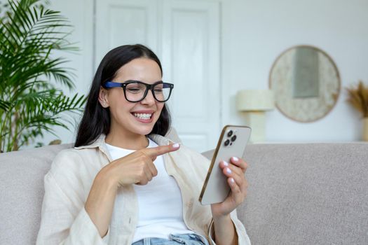 Happy young student girl in glasses sitting on sofa at home, holding phone, chatting and chatting with friends, smiling.