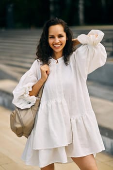 Smiling curly brunette girl in a white dress with a backpack putting her hand behind her head. High quality photo