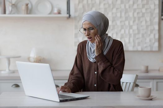 Muslim woman toothache at home, business woman in hijab working remotely from home, sitting in the kitchen using a laptop.