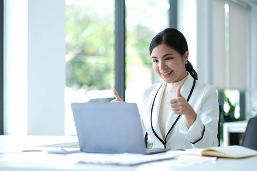 Distant communication. Friendly pretty asian brunette girl, manager, freelancer, sit in the kitchen, talking on video call by laptop with the client or employees, discussing project, smiling.