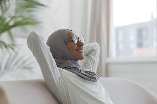 Young curvy muslim woman in hijab and glasses resting at home, woman on sofa with arms behind head, smiling with closed eyes