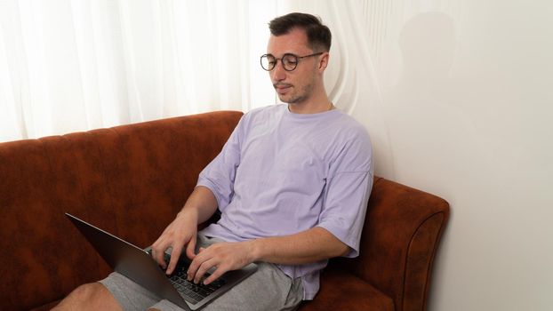 A man with glasses types on a laptop on the couch of the house. High quality photo