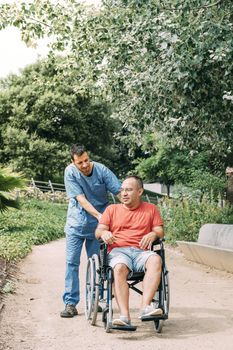 disabled man in wheelchair talking during a walk with his nurse at park, concept of medical care and rehabilitation of people with disabilities and reduced mobility problems, vertical photo