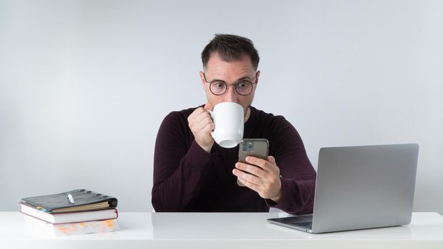 A man at work drinks coffee and looks at the phone screen in surprise with big eyes - unexpected news. High quality photo