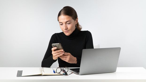 A girl types a message on her phone at her desk with a laptop and notepad. High quality photo