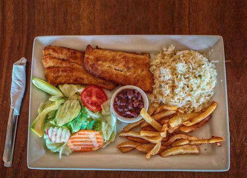 Plate of fried fish fillet with salad and french fries on wooden table, Top view of delicious fried fish fillet with french fries, rice and salad served on wooden table. Gourmet food plate