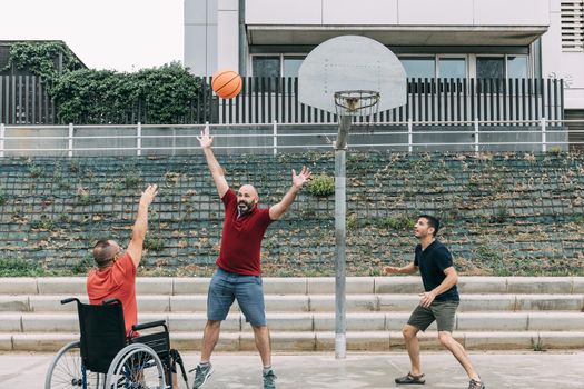 joyful disabled man in wheelchair throwing the ball to basket with friends, concept of adaptive sports and physical activity, rehabilitation for people with physical disabilities
