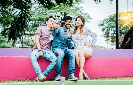 Lifestyle of three friends sitting in a park taking a selfie, Three teenage friends sitting in a park taking selfies. Meeting of three happy friends taking a selfie sitting in the park