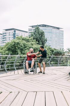 disabled man in wheelchair shake hands with his friend during a walk at park at city, concept of friendship and integration of people with disabilities and reduced mobility problems, vertical photo