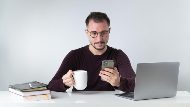 A man with glasses with a phone in his hand and a cup of coffee on his desk with a laptop. High quality photo