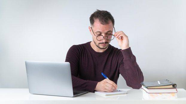 A man with glasses works at home or in the office with a laptop, makes a note in a notebook. High quality photo