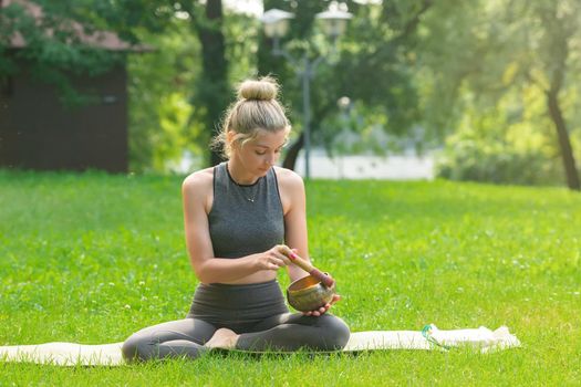 A beautiful woman in a gray top and leggings, sitting on the green grass in the park in summer, holds a meditation bowl, drives a stick on it. Copy space