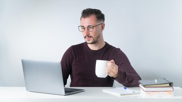 A man with glasses with a cup of coffee works at a laptop at the workplace. High quality photo