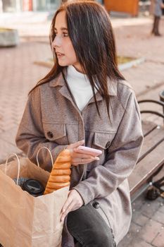 A stylish brunette woman walks around the autumn city. The brunette is sitting on a bench outside with a glass of coffee and a phone, holding a craft bag with a baguette.