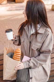 A stylish brunette woman walks around the autumn city. The brunette is sitting on a bench outside with a glass of coffee and a phone, holding a craft bag with a baguette.