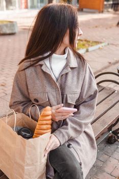 A stylish brunette woman walks around the autumn city. The brunette is sitting on a bench outside with a glass of coffee and a phone, holding a craft bag with a baguette.