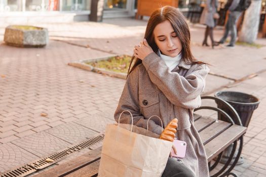 A stylish brunette woman walks around the autumn city. The brunette is sitting on a bench outside with a glass of coffee and a phone, holding a craft bag with a baguette.
