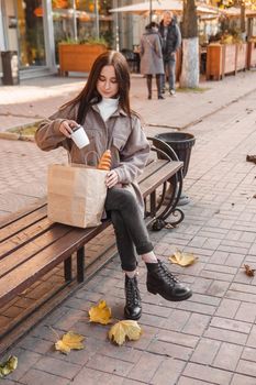 A stylish brunette woman walks around the autumn city. The brunette is sitting on a bench outside with a glass of coffee and a phone, holding a craft bag with a baguette.