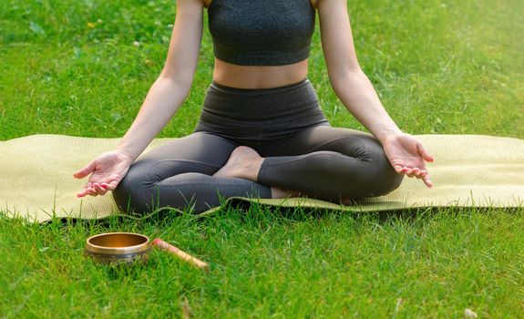 A slender woman in a gray top and leggings, sitting on the green grass in summer, meditates, there is a meditation bowl on the grass next to her. Copy space