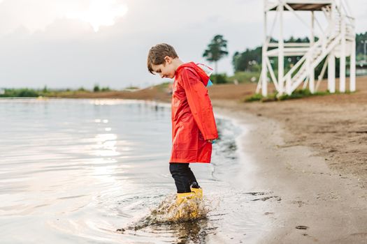 Boy in a red raincoat and yellow rubber boots playing with water at the beach. School kid in a waterproof coat jumping in water at sea. Child having fun with waves at the shore.