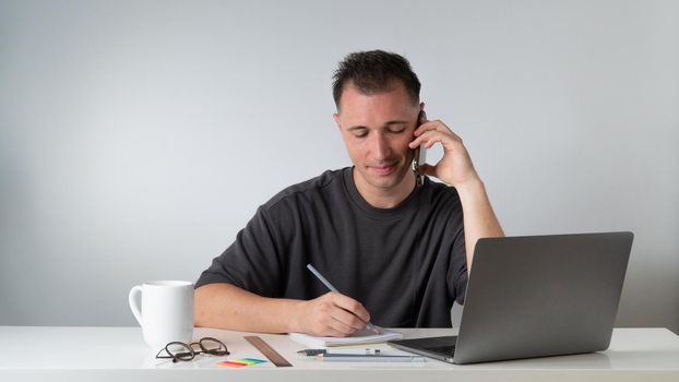 Male student writes a lecture in a notebook and talks on the phone with a laptop. High quality photo