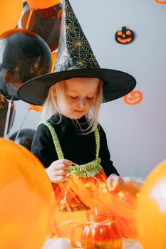 Children's Halloween - a girl in a witch hat and a carnival costume with airy orange and black balloons at home. Ready to celebrate Halloween.