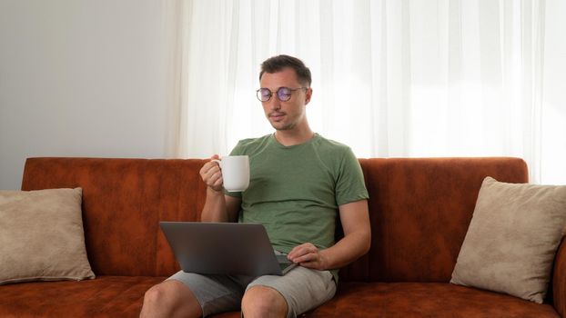 A man with a laptop sits on the couch at home and drinks coffee. High quality photo