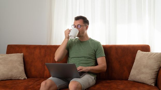 A man works at the computer on the couch and drinks coffee. High quality photo