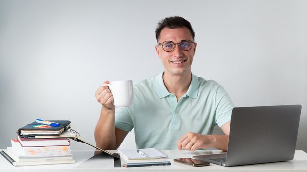 A student guy drinks coffee at a table with textbooks, books, a laptop. High quality photo