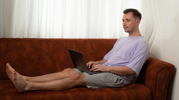 A man lies on the couch and types in a laptop, working at the computer at home. High quality photo