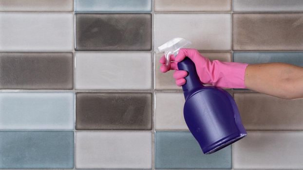 A woman in rubber gloves washes tiles in the kitchen or bathroom with a sponge and sprays detergent
