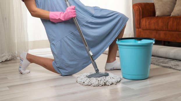 A woman with a mop and a bucket in pink rubber gloves on her knees washes the floor