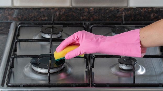 A woman's hand in a pink rubber glove washes a gas stove with a sponge
