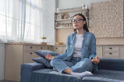 Young beautiful Asian girl meditates at home sitting on the couch, resting during the day, sitting in the lotus position