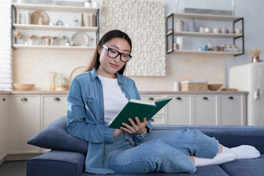 Young beautiful asian woman in glasses and denim shirt studying at home, happy female student smiling reading book sitting on sofa in living room