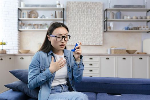 Young beautiful woman sick at home, having asthma and difficulty breathing, asian woman wearing glasses sitting on sofa in living room uses inhaler to ease breathing