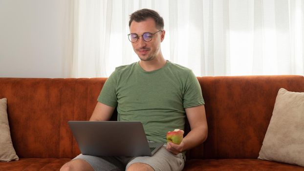 A man with a laptop on the couch eats an apple. High quality photo