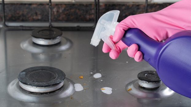 A woman's gloved hand sprays detergent on a dirty gas stove. High quality photo