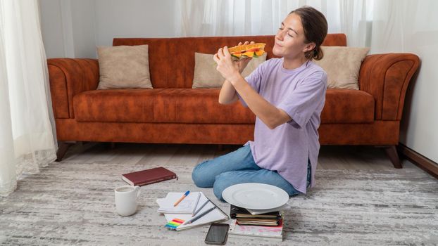 A hungry female student eats a sandwich and prepares for an exam at home. High quality photo