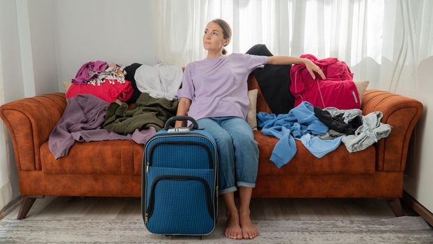 A woman among a pile of clothes with a suitcase sits on the couch. High quality photo