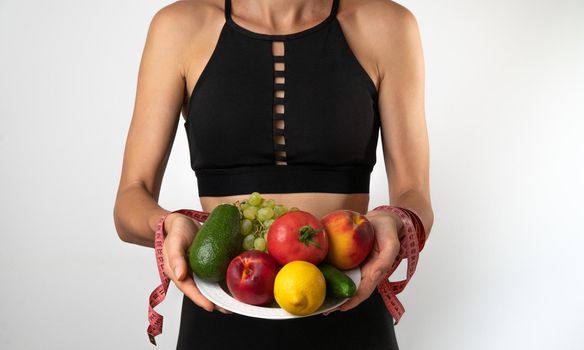 A woman in a sports uniform holds a plate with fruits and vegetables and measuring tape in her hands, trainer nutritionist. High quality photo