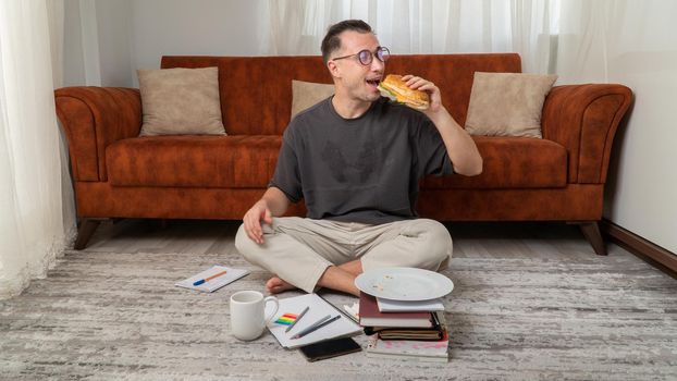 A student guy eats a sandwich while studying at home. High quality photo