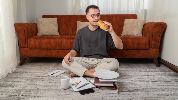 Hungry male student eats a sandwich behind textbooks at home. High quality photo