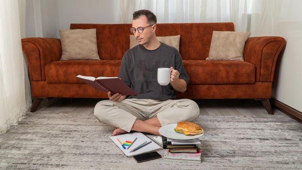 Male student drinks coffee and reads a book, studies at home, snacks. High quality photo