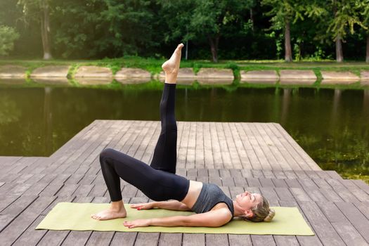 A woman in a gray top and leggings, on a wooden platform by a pond in a park in summer, does yoga, performing a pose with her leg raised up. Copy space