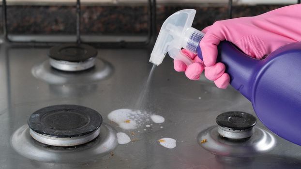 A hand in a pink glove sprays detergent on a dirty gas stove for cleaning. High quality photo